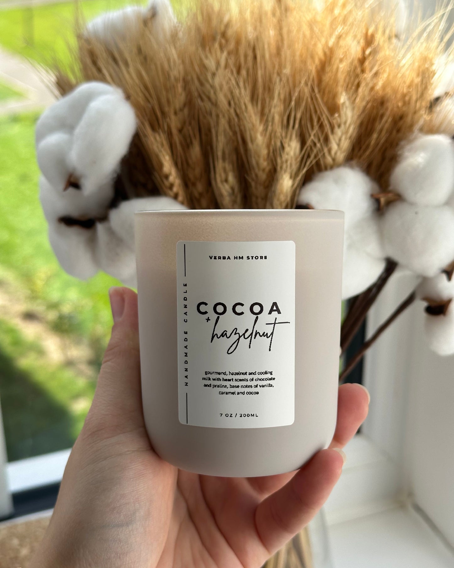 Candle labeled 'Cocoa Hazelnut' held in a hand with a blurred natural background
