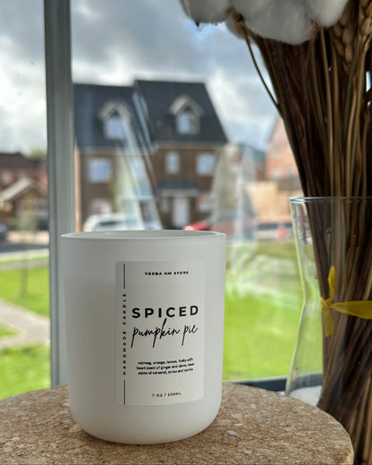 White candle labeled 'Spiced Pumpkin Pie' on a windowsill with a view of houses outside.