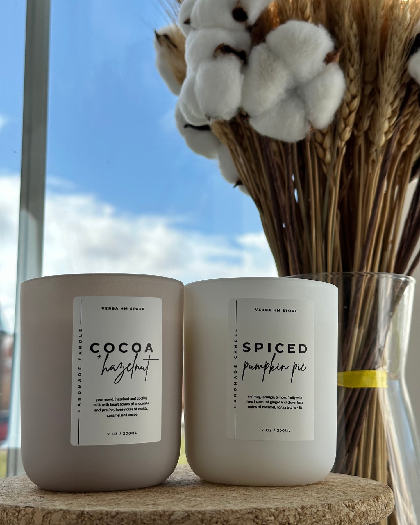 Two candles with labels 'Cocoa Hazelnut' and 'Spiced Pumpkin Pie' on a surface with a decorative background.